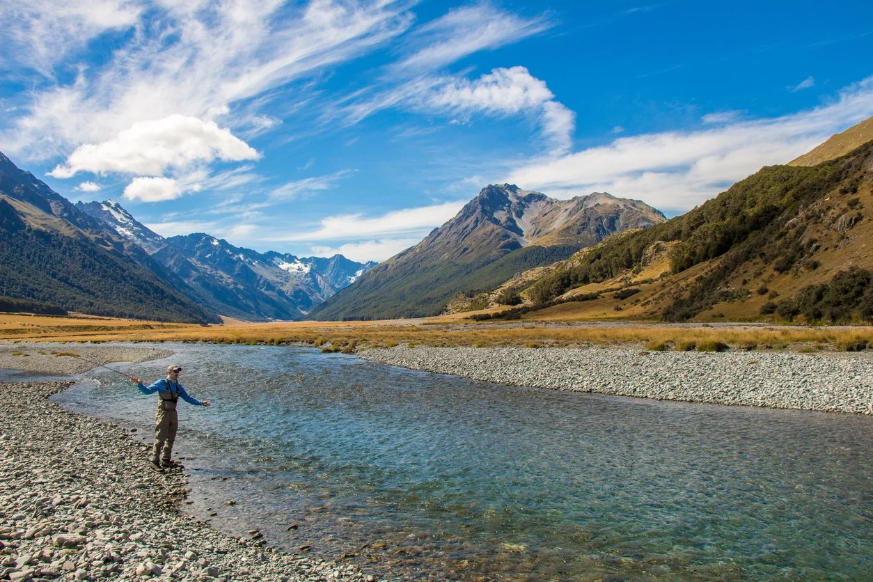 Person fishing in a river surrounded by mountains and blue sky with clouds.