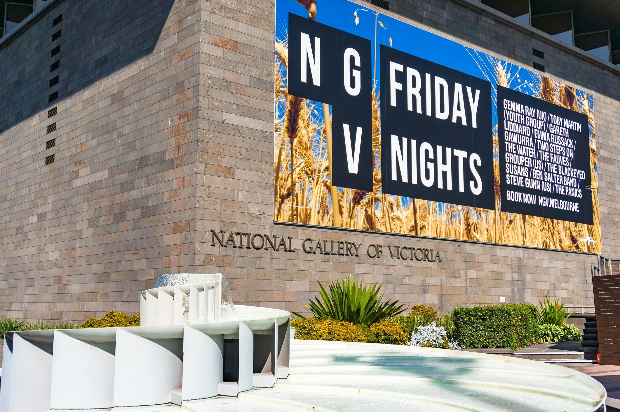 The National Gallery of Victoria in Melbourne, Australia, displays a banner for Friday Nights at the Gallery, with its iconic fountain flowing under a bright, sunny sky.