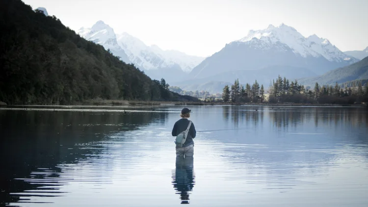 A person stands in a calm lake, fishing, with snow-capped mountains and a forested landscape in the background.