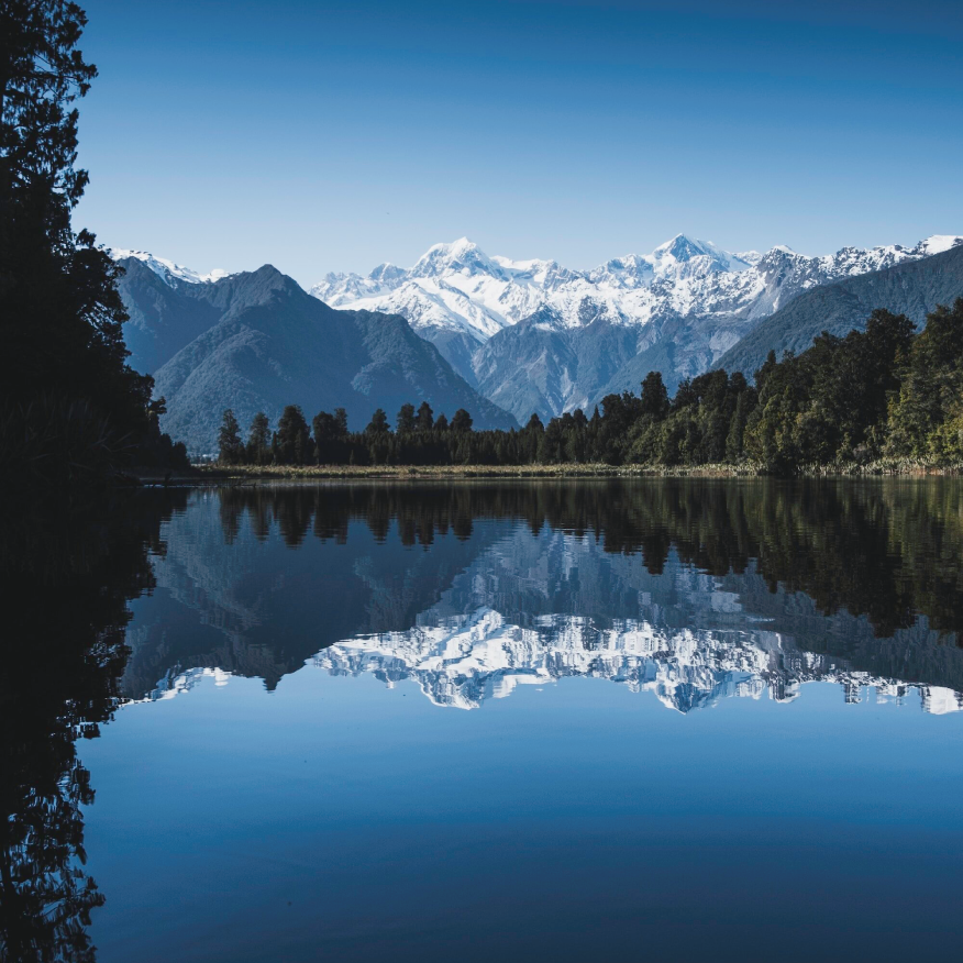 Snow-capped mountains reflected in a calm lake with forested shoreline under clear blue sky.