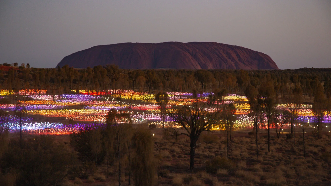 The Field of Light installation at Uluru, Australia, features a nighttime display of colorful, illuminated glass spheres across the desert landscape, with Uluru silhouetted in the background under the night sky