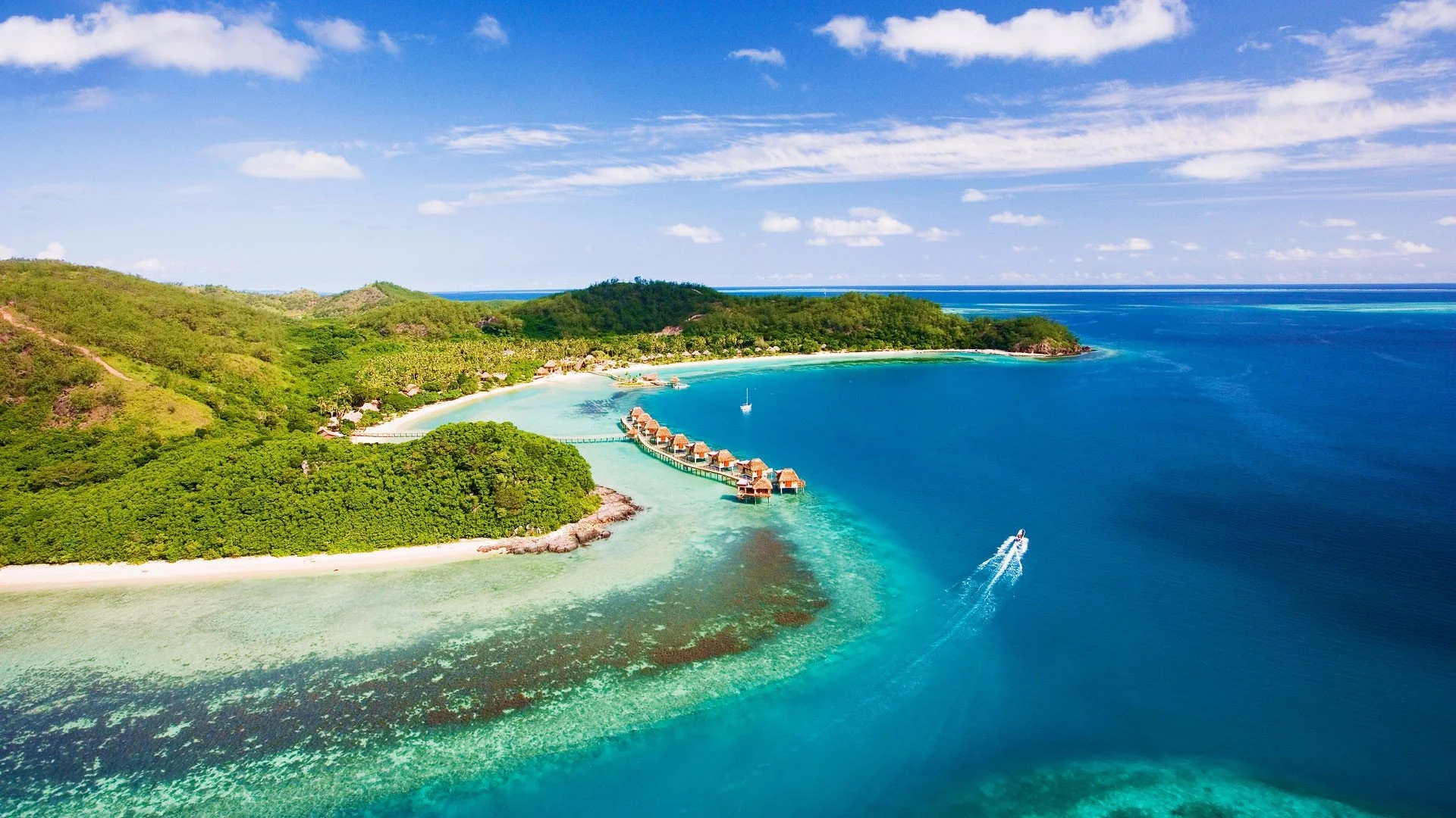 Aerial view of a Fiji island with a fringing reef, turquoise lagoon, a small cluster of overwater bures, and a boat crossing the water.