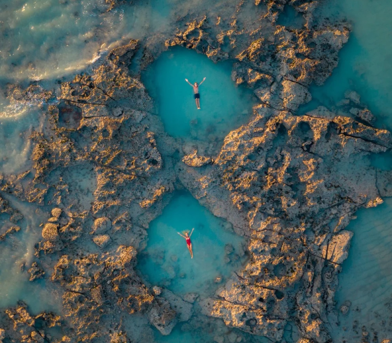 An aerial view of a couple each swimming separately in natural rock pools at Gantheaume Point, surrounded by red sandstone cliffs and clear blue waters in Broome, Western Australia.