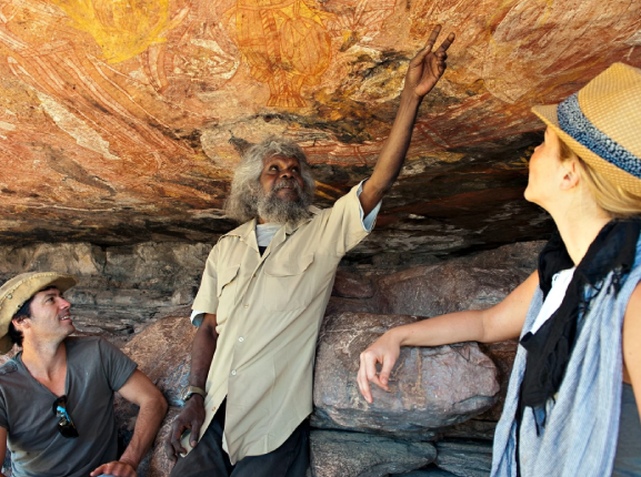 Three people in a cave observing ancient Aboriginal rock art painted on the ceiling. One man with gray hair and beard points at the artwork while two others, a woman and a man, look on.