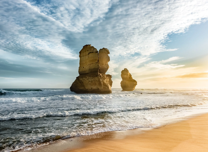 Two large sea stacks on a sandy beach at sunset with waves crashing, cloudy sky, and sunlight on the horizon.