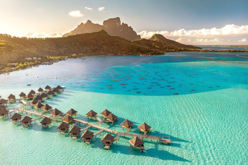 Overwater bungalows with thatched roofs resting on stilts in turquoise water near a green hillside and mountain range under a partly cloudy sky.