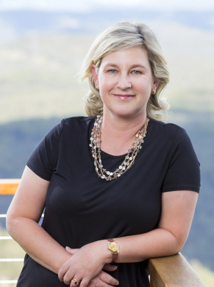 A smiling woman with blonde hair, wearing a black top, layered necklaces, a gold watch, and rings, leaning on a wooden railing outdoors with a blurred scenic landscape in the background.