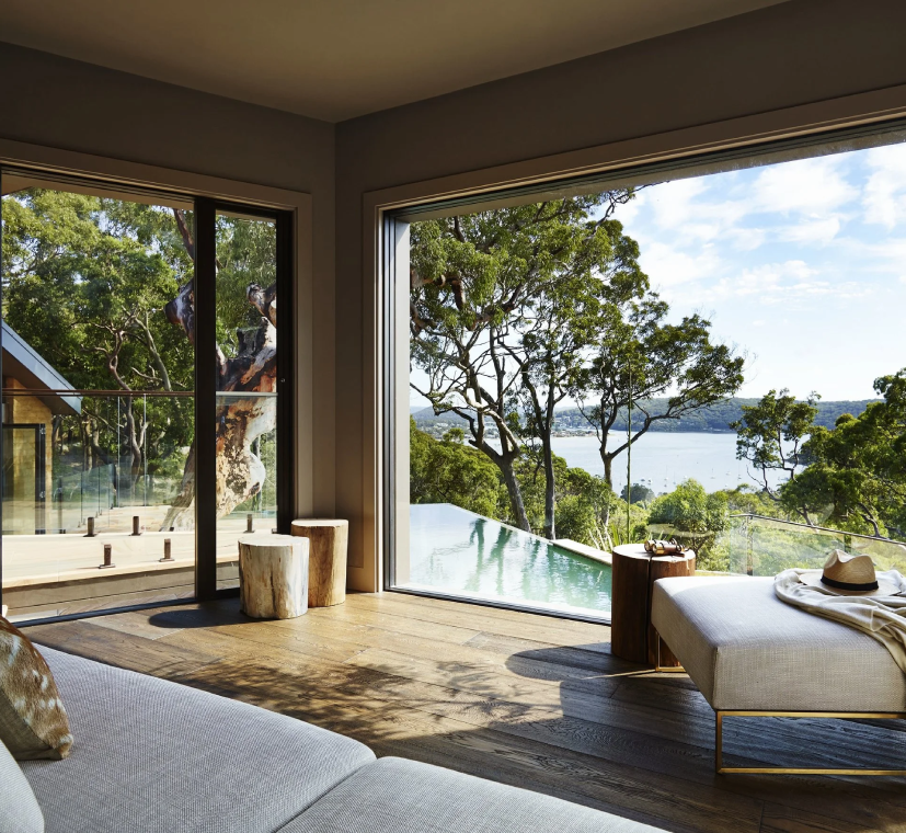 Living room with large windows overlooking trees and a body of water, with a white chaise lounge, a hat and blanket, wooden stools, and an indoor-outdoor feel.
