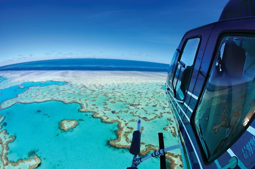 Aerial view of a helicopter flying over a turquoise reef and ocean with a clear blue sky.