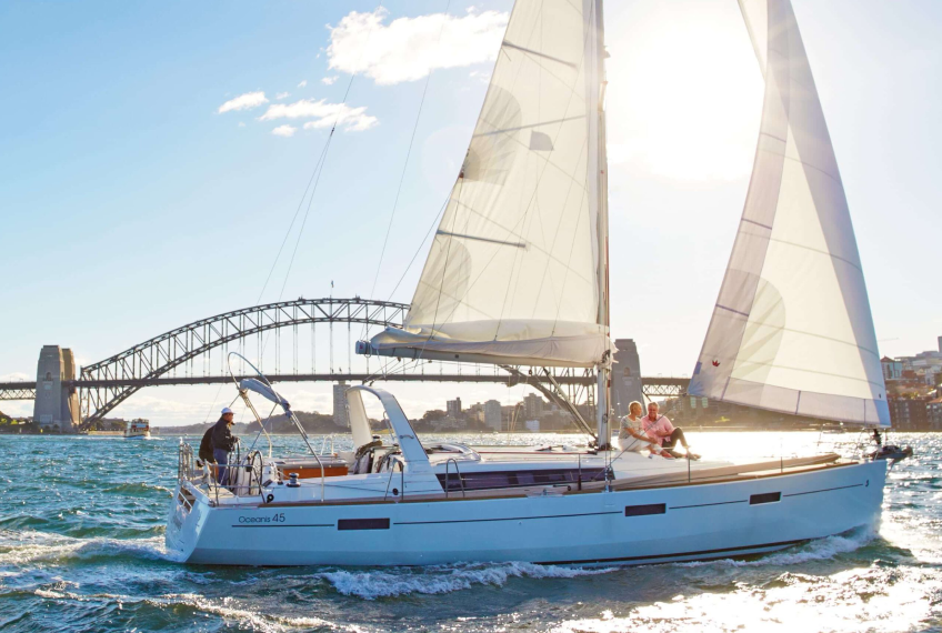 A sailboat with two people aboard sailing in a city harbor with a bridge in the background, and the sun shining brightly.