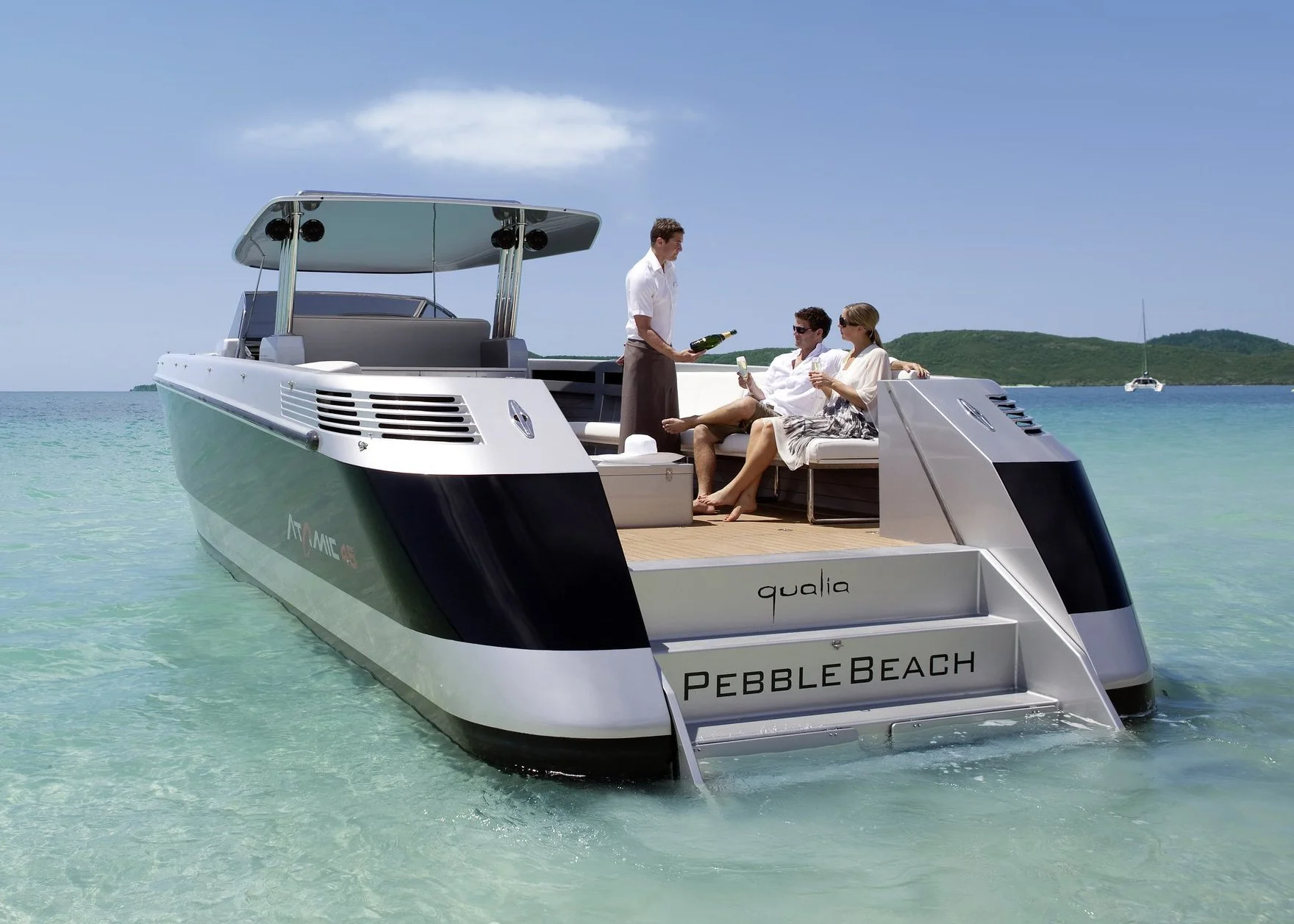 People enjoying a luxurious boat on clear, turquoise water with a small island and a sailboat in the background.