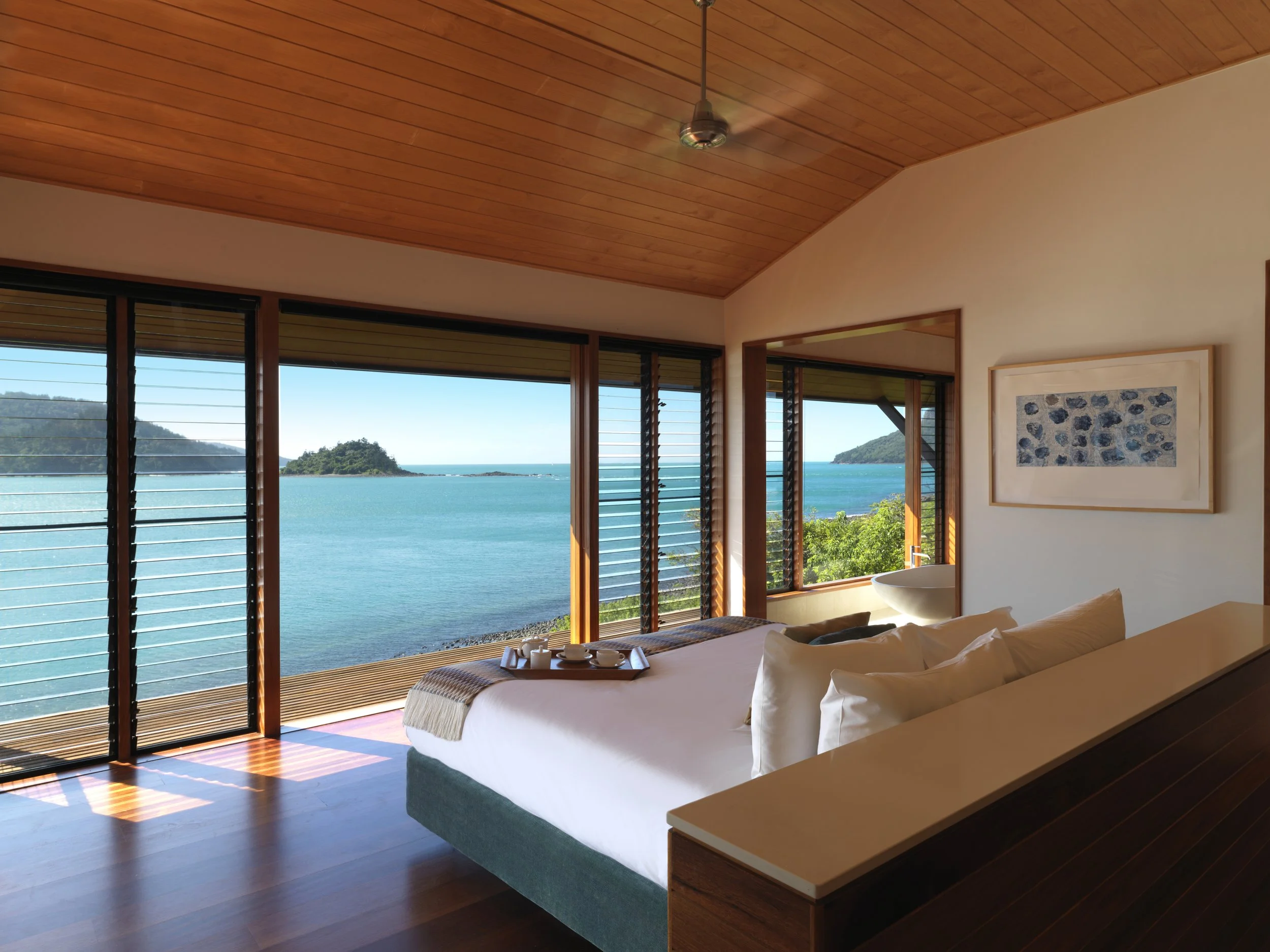 Room with large windows overlooking a blue ocean and islands, wooden ceiling, bed with white linens and pillows, and wall art.