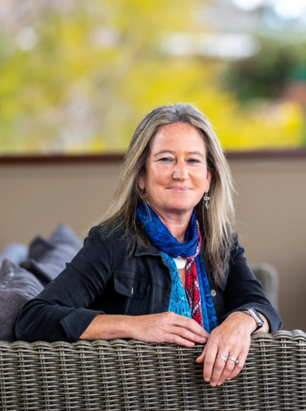 A woman with long blonde hair sits at a wicker outdoor patio table, wearing a black jacket and a blue scarf with a red pattern, against a blurred background of green trees and a beige wall.