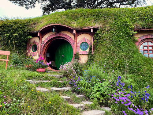 A Hobbit-style house built into a grassy hillside with a round green door, small circular windows, colorful flowers, and stone steps leading up to the entrance.