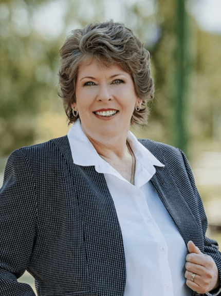 A smiling woman in business attire standing outdoors, wearing a dark patterned blazer over a white shirt, with a blurred natural background.