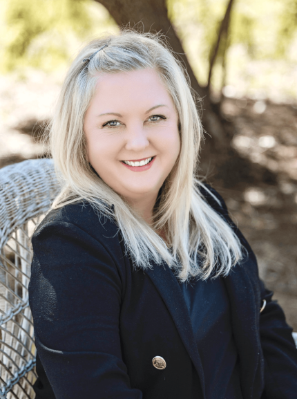A smiling woman with long blonde hair and blue eyes, wearing a black blazer, sitting outdoors on a white wicker chair with a blurred outdoor background.