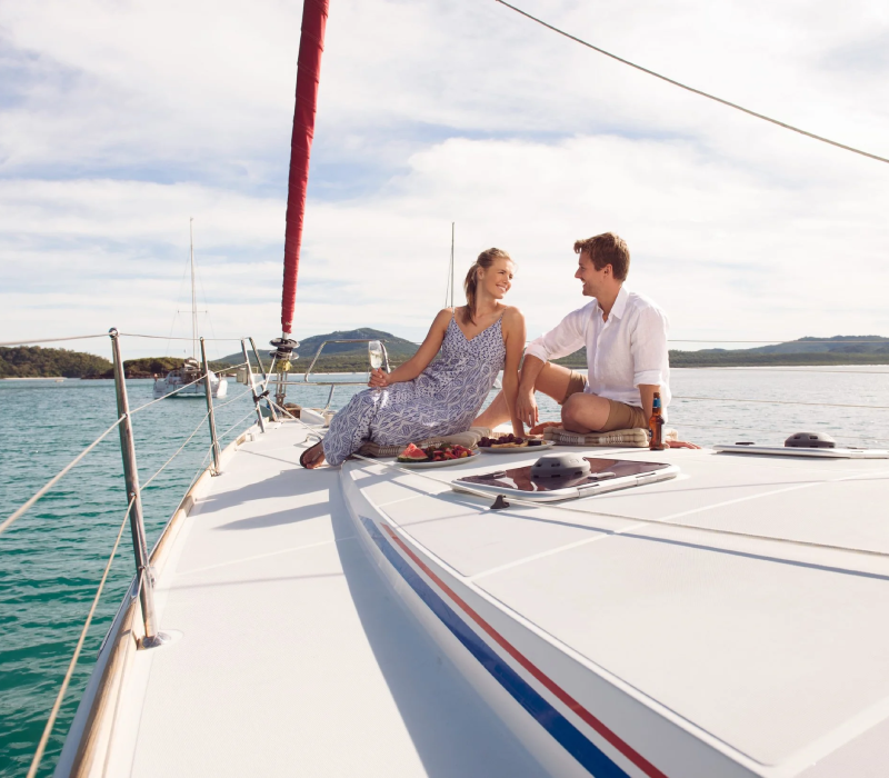 A couple enjoying a picnic on a sailboat with scenic water and islands in the background.