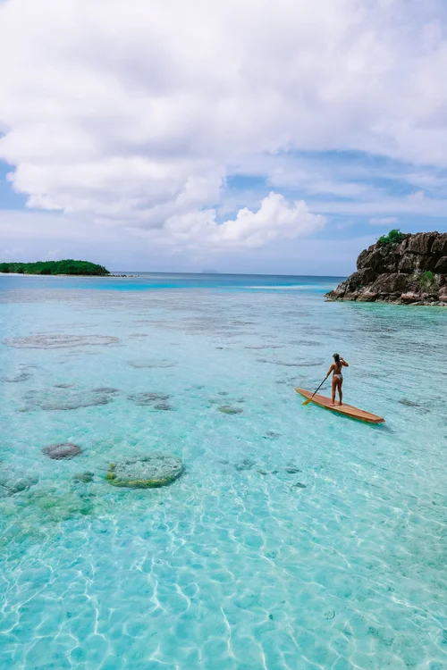 A person standing on a stand-up paddleboard in clear, shallow turquoise water near a rocky coastline with green trees, under a partly cloudy sky.