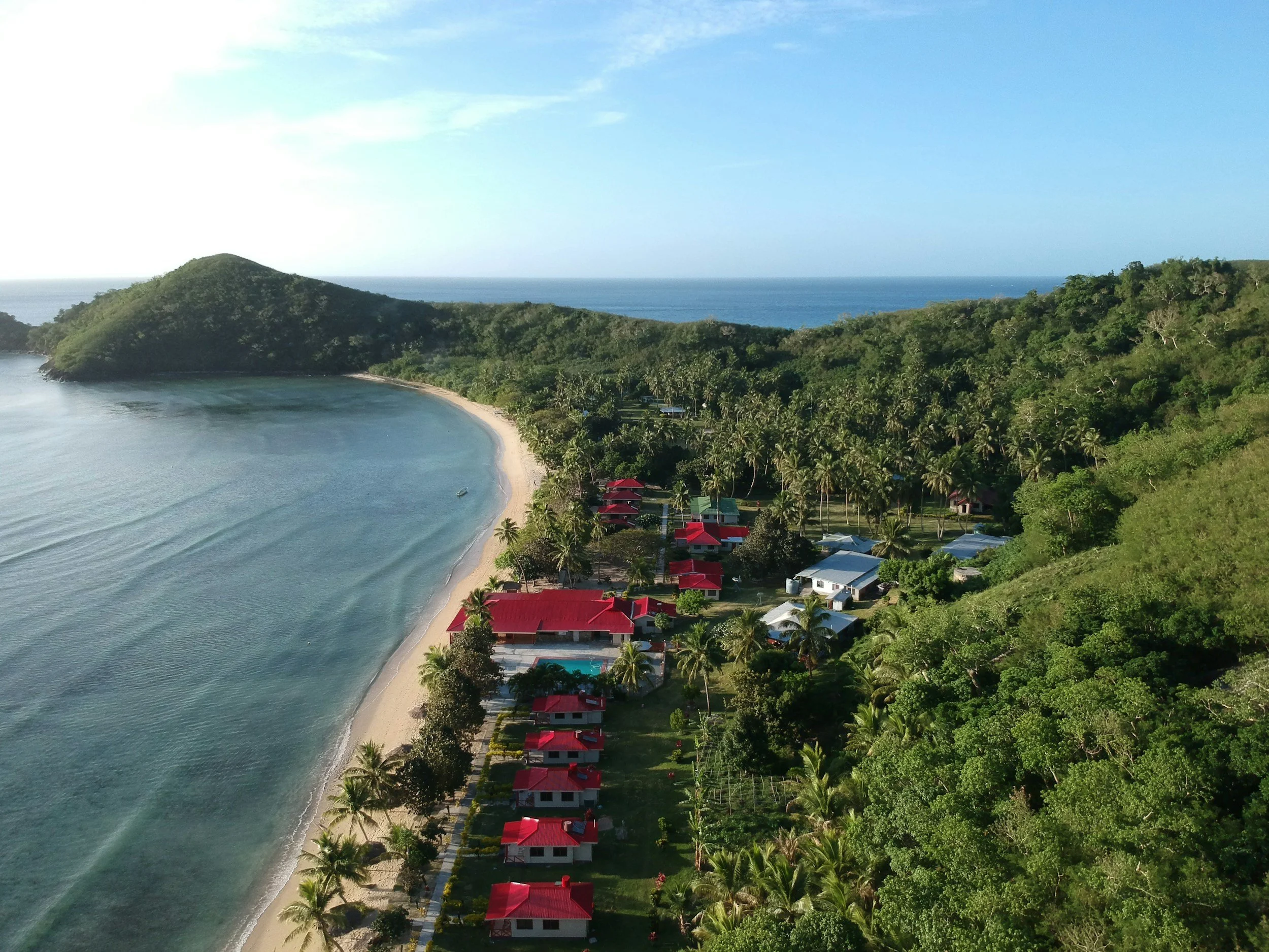 Aerial view of a tropical beach with clear water, a sandy shoreline, and a lush green hillside with trees and houses, some with red roofs, and a distant view of the ocean and blue sky.