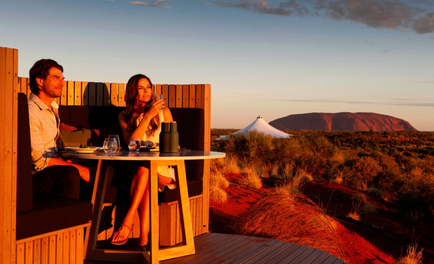A man and woman dining on a wooden deck with desert landscape and mountains in the background at sunset.
