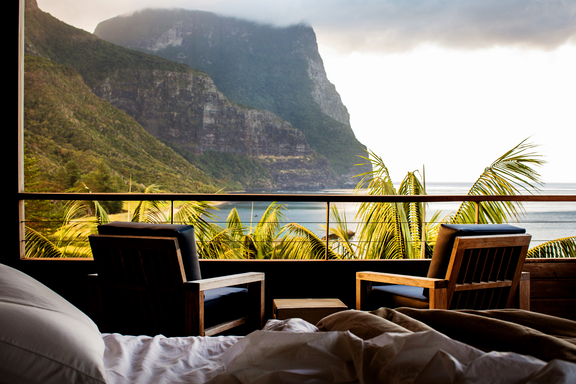 Photo of Capella Lodge bedroom view. In the foreground, there are palm trees, and in the background the open ocean and mountains.