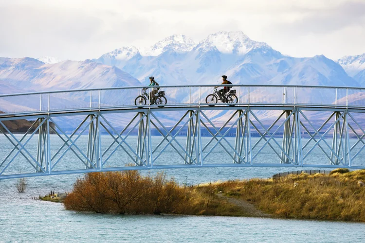 Two cyclists cross the iconic footbridge over the turquoise waters of Lake Tekapo, New Zealand, with snow-capped mountains in the background