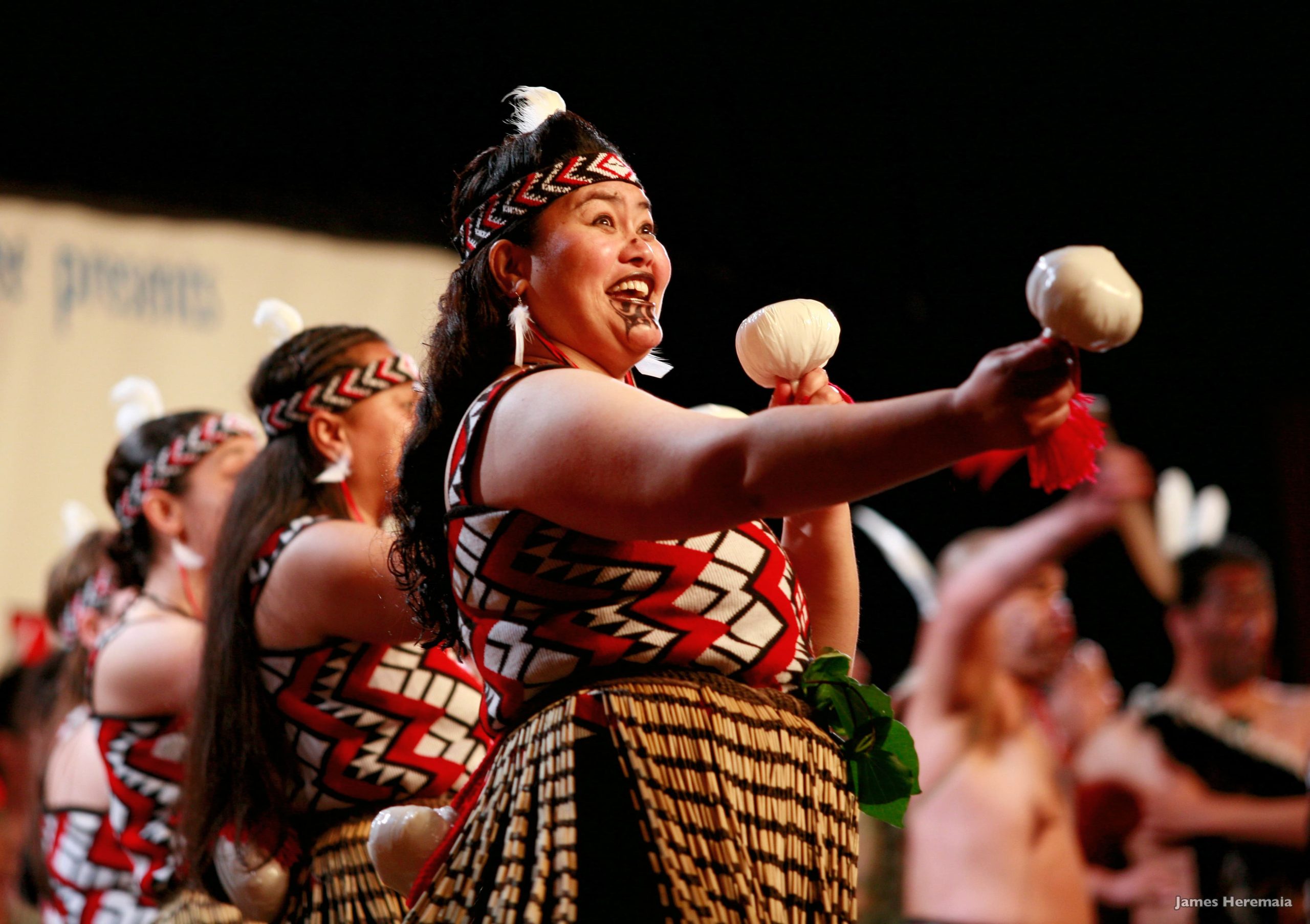 A group of Maori women share poi dance, a skillful dance that involves control of ball swung on string. They are wearing traditional clothing and smiling.