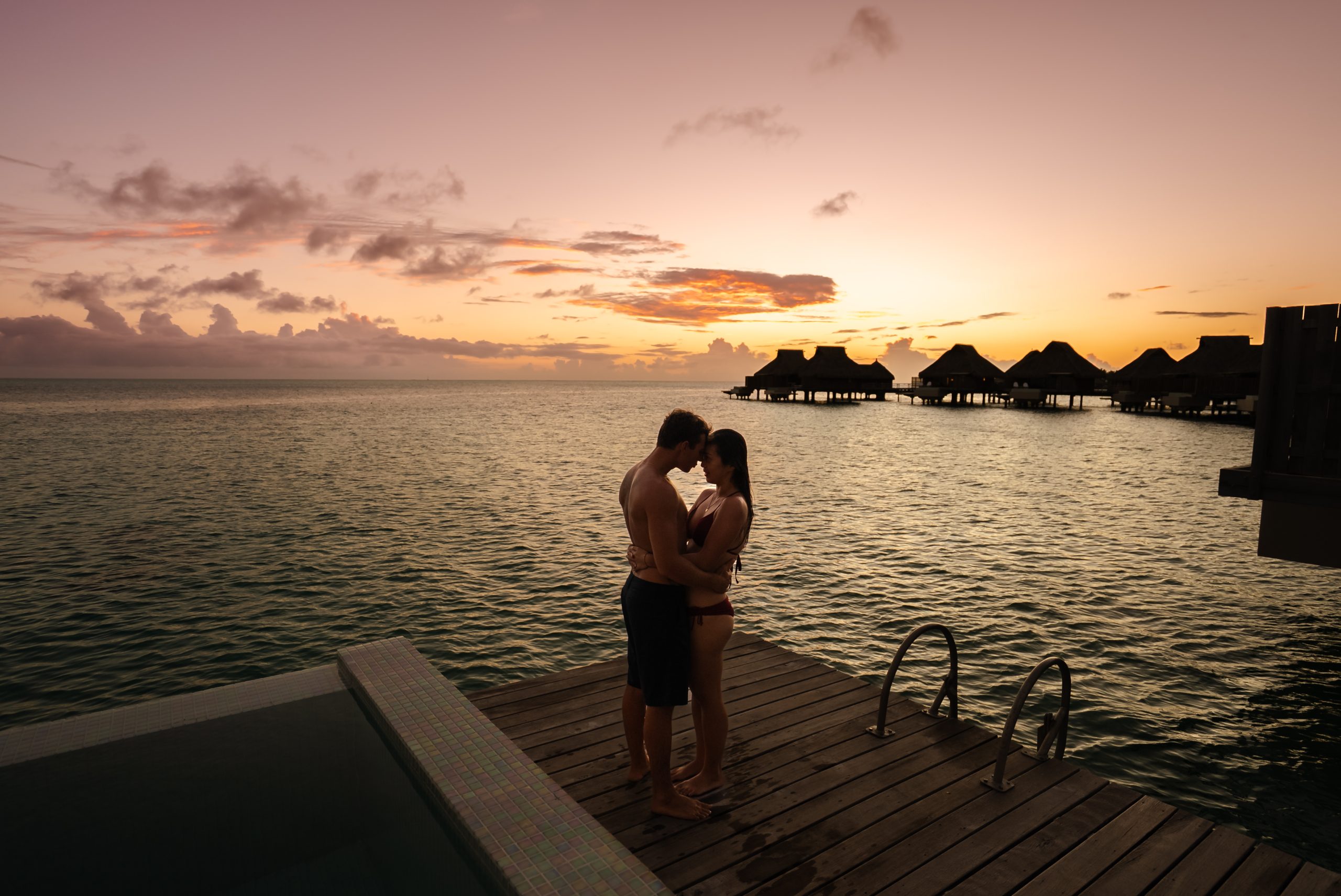  A couple stands nose to nose, happy on vacation together. In the background, there are overwater bungalows and a sunset over the ocean.
