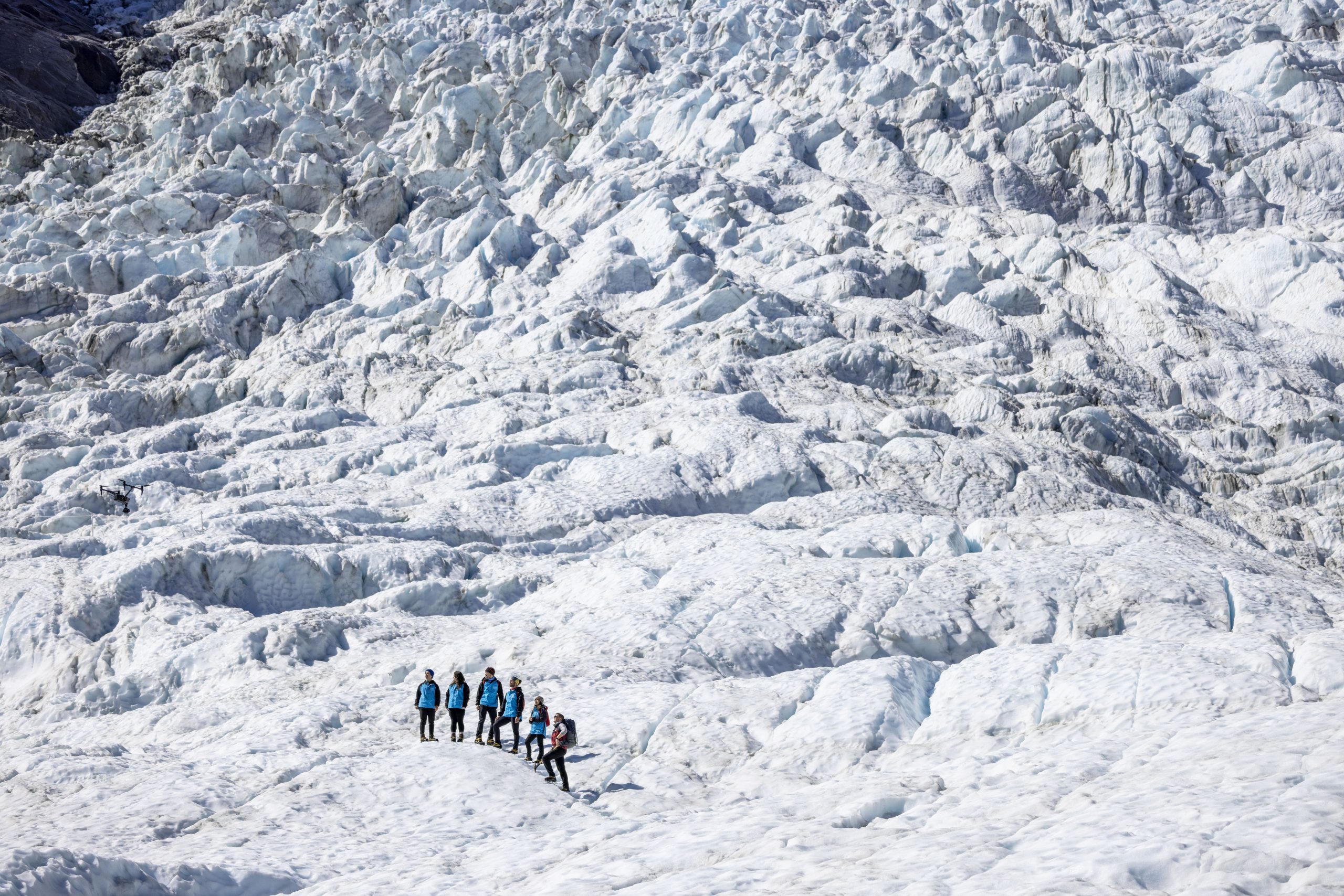 A group of hikers takes a guided trek through Fox Glacier, NZ. Aerial view, landscape covered in bright, white snow.