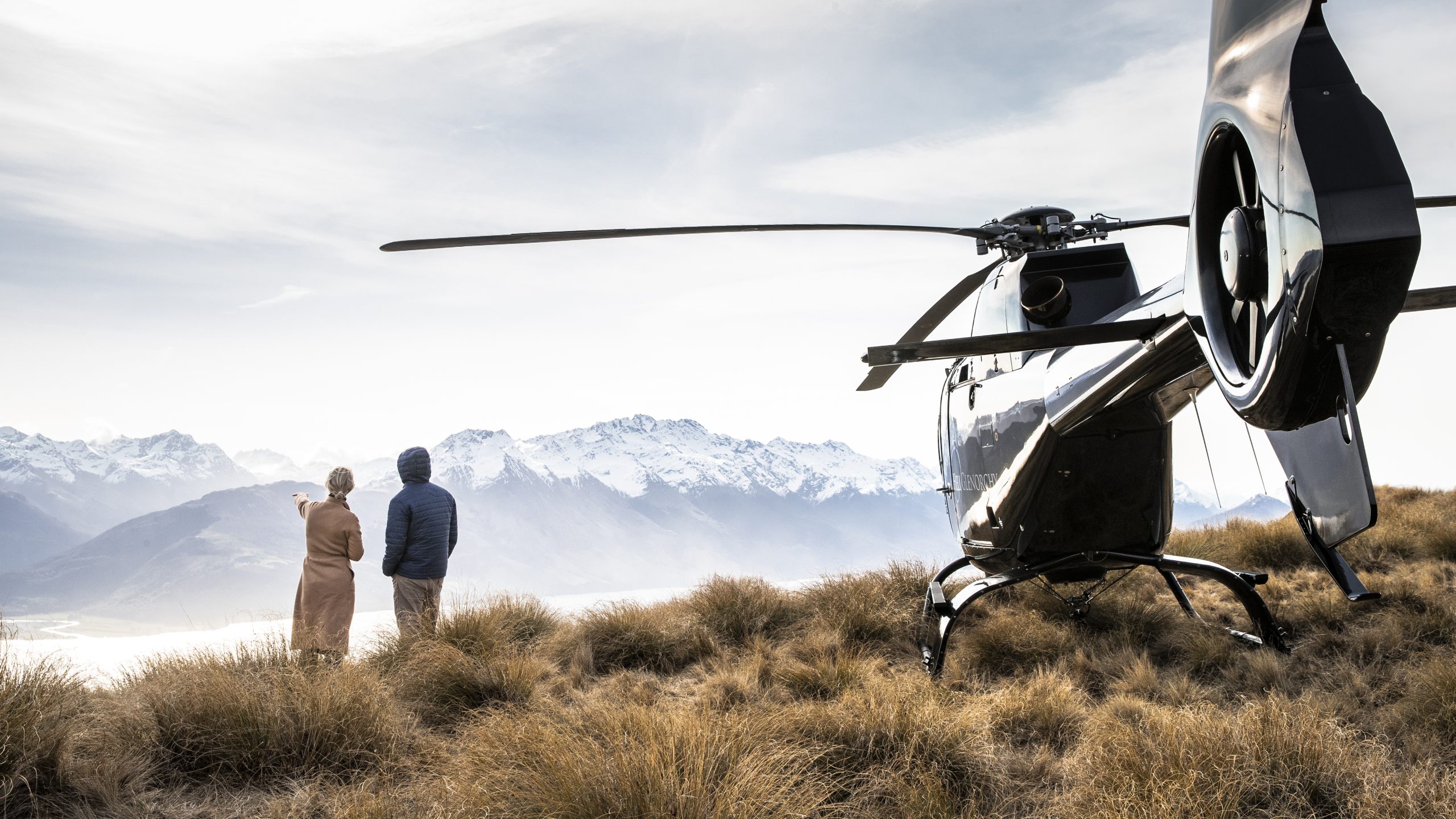A couple looking out at snow-capped mountains next to a black helicopter parked on a grassy alpine ridge.