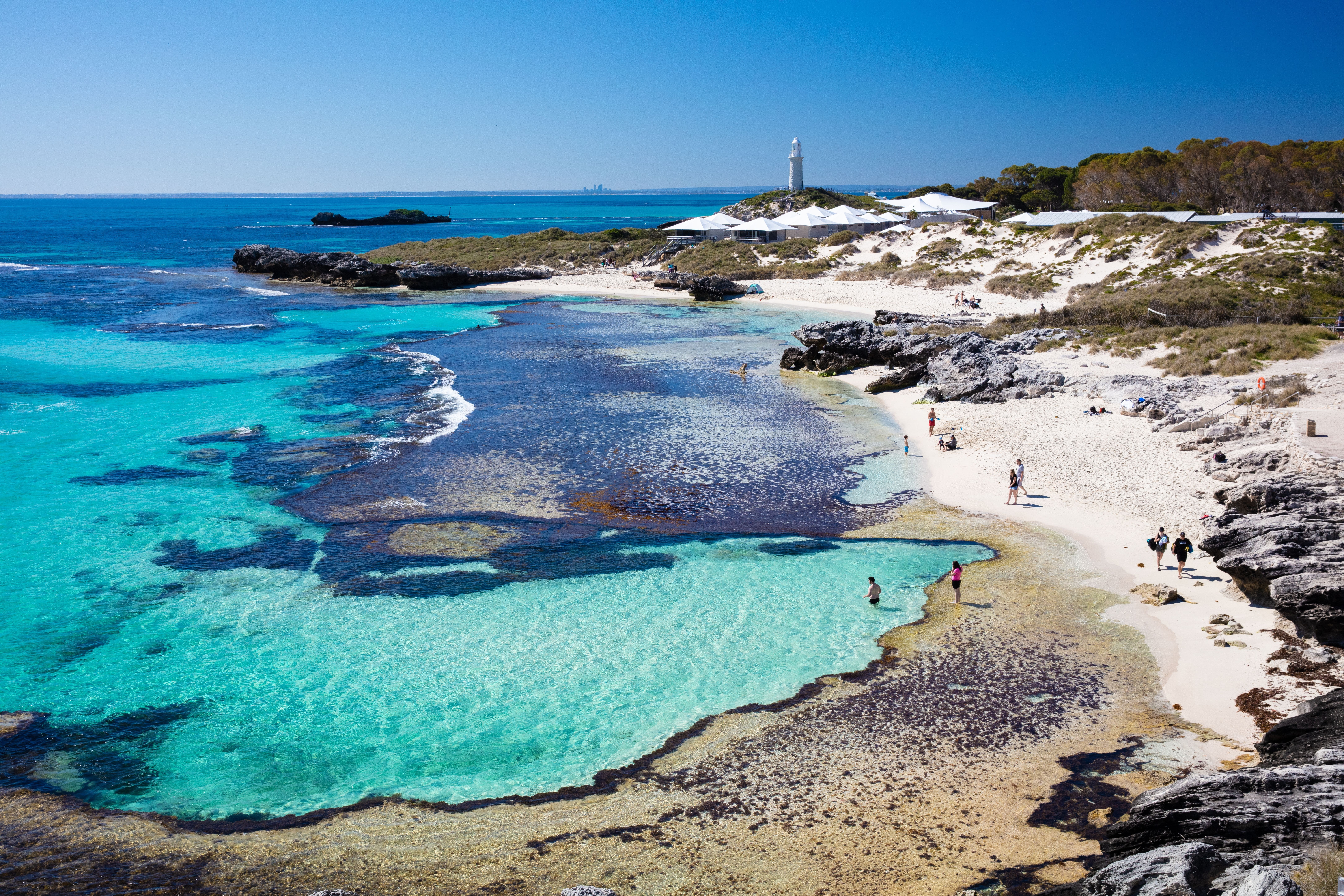 Ariel view of the Basin on Rottnest Island. Beachgoers wade in the crystal clear waters and a lighthouse is seen in the distance. 