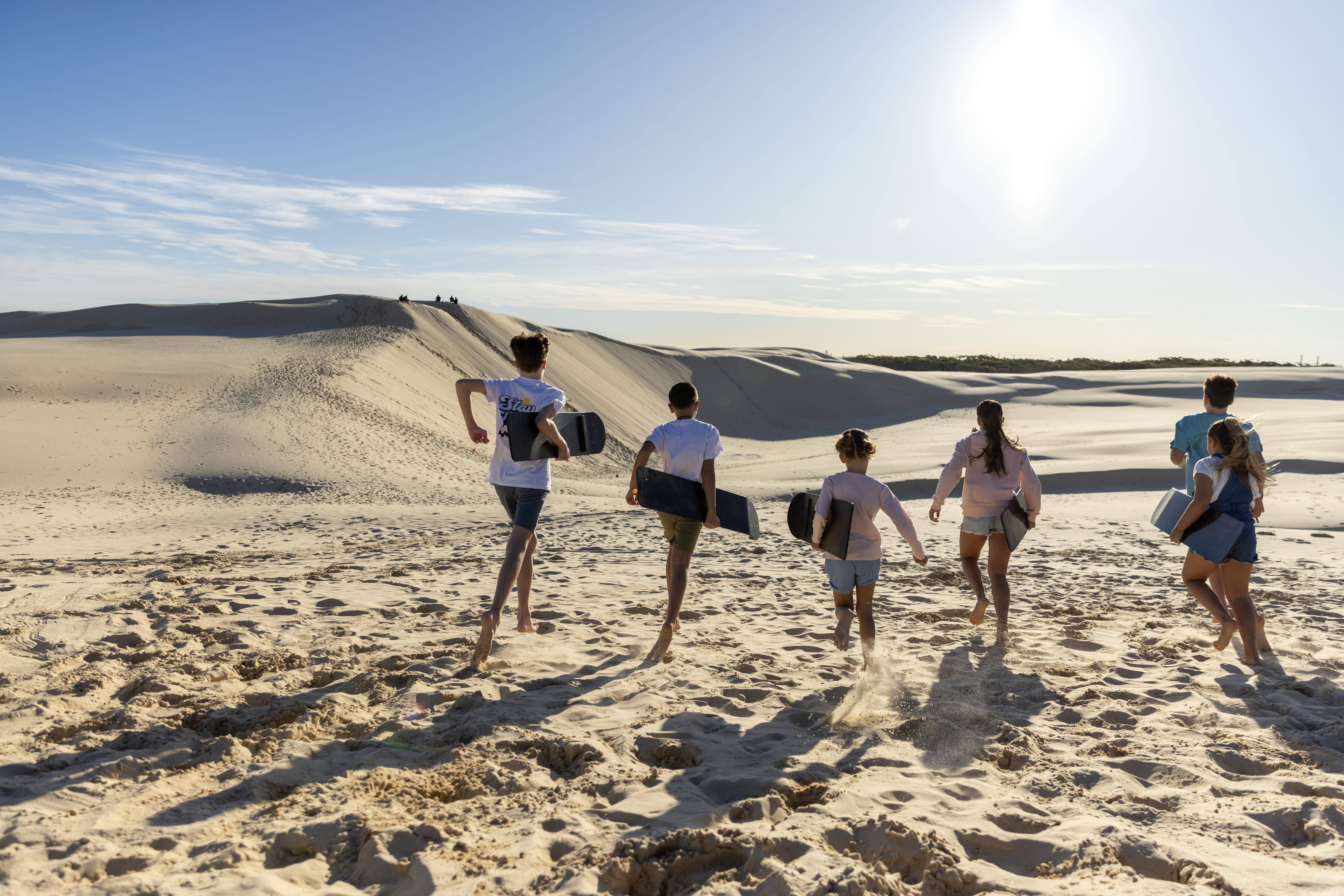 Family exploring the sand dunes at Stockton Beach, running the opposite direction with skim boards in hand.