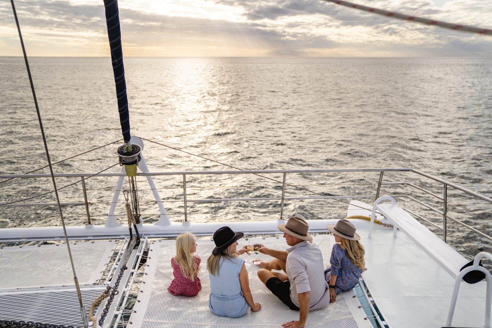 Family sits aboard private charter on Ningaloo Reef, Coral Coast. The sun sets in the background and the parents clink wine glasses.