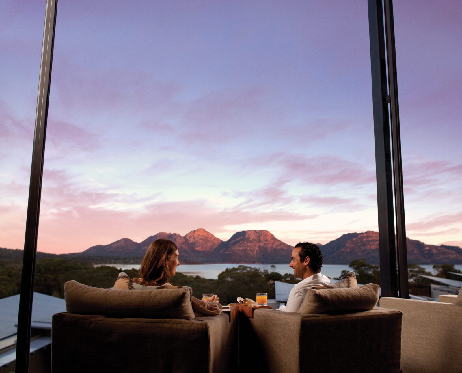 A couple relaxes in armchairs by a floor-to-ceiling window, watching sunset light over red mountains and a still bay.