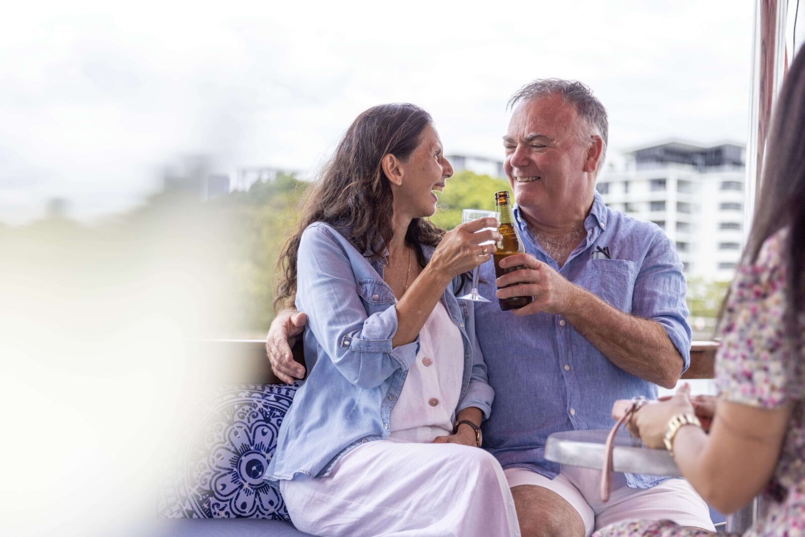 A senior couple shares a toast together on a riverside terrace along the Brisbane River, with the city skyline behind them.