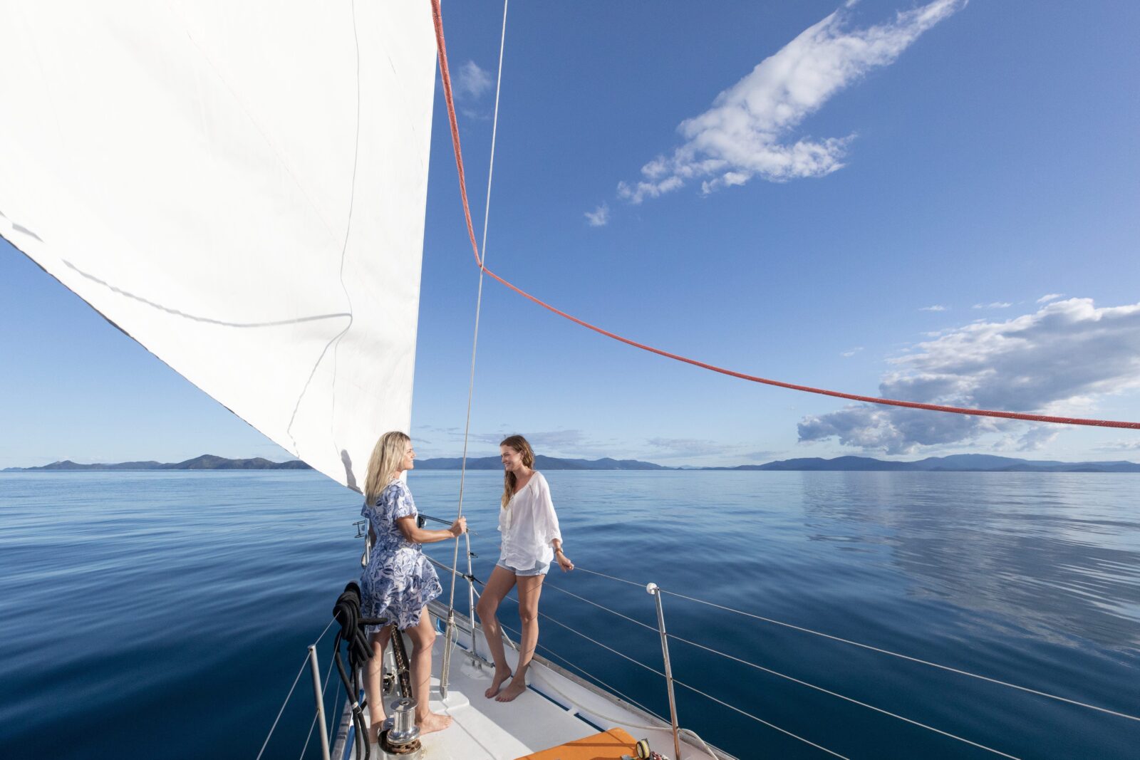 Two women stand on the deck of a sailing yacht on calm blue water under a partly cloudy sky.