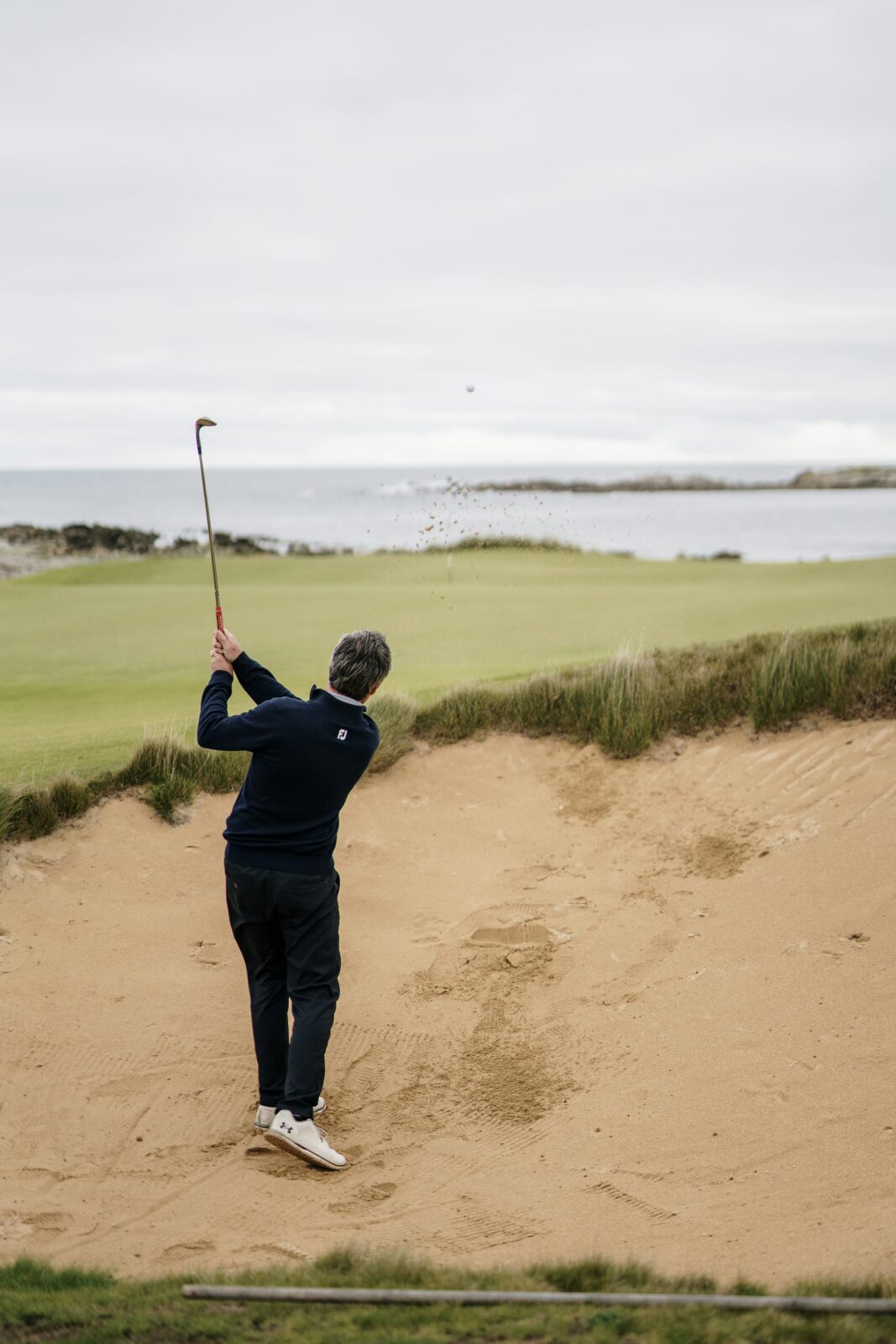 A golfer plays a bunker shot on a seaside golf course in Australia, with the ocean and coastline in the background.