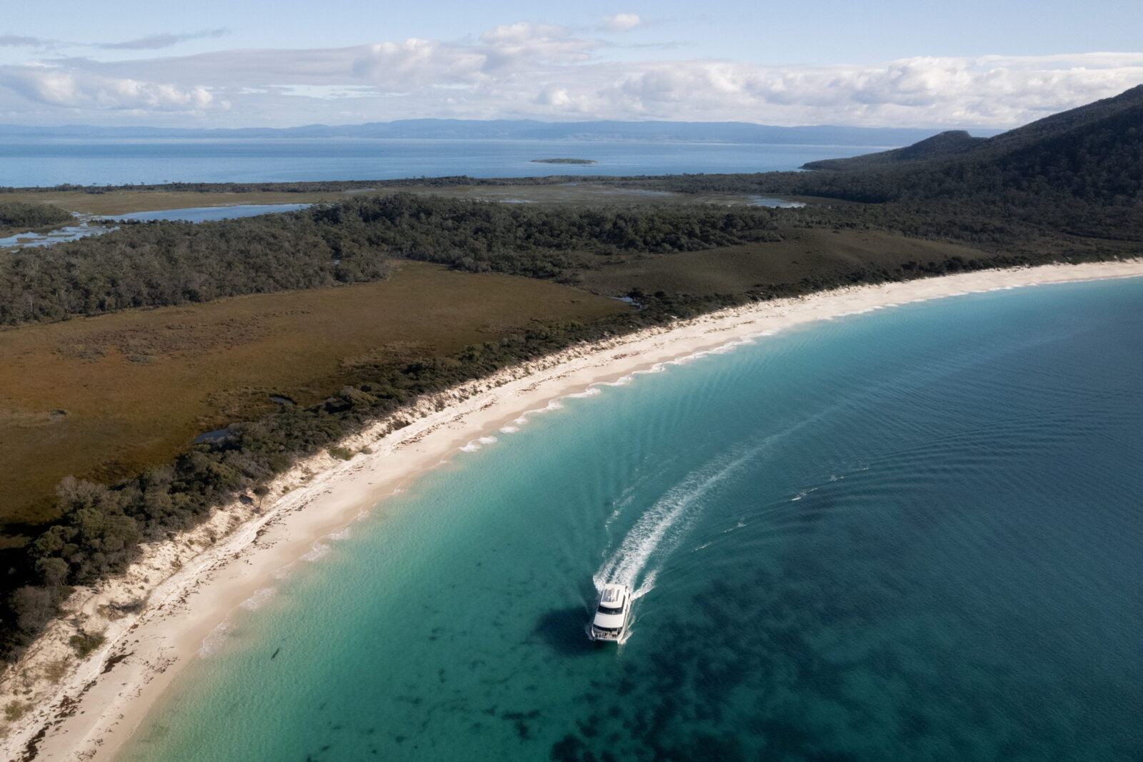 Aerial view of a boat cruising into a secluded Tasmanian beach cove with clear turquoise water.