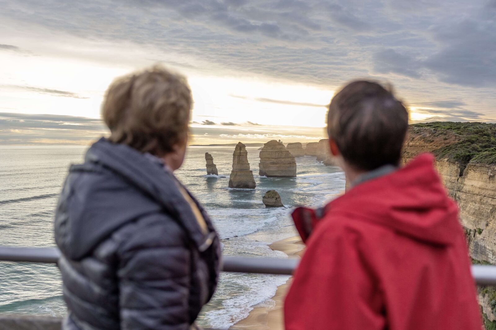 Two senior travellers stand at a coastal lookout, taking in ocean views along Australia’s coastline at sunset.