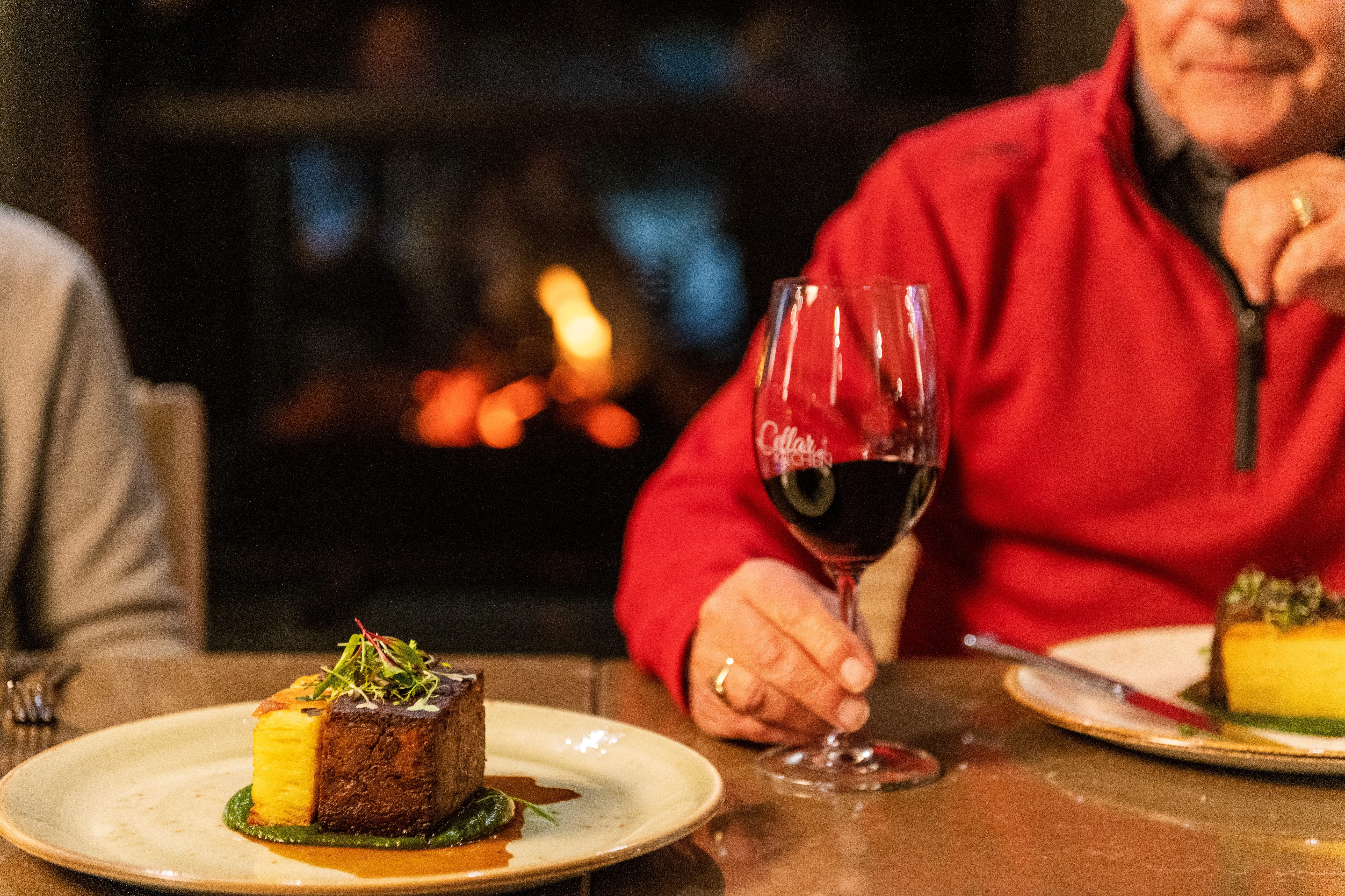 An older man in a red sweater raises a glass of red wine at a restaurant table set with a plated dish.