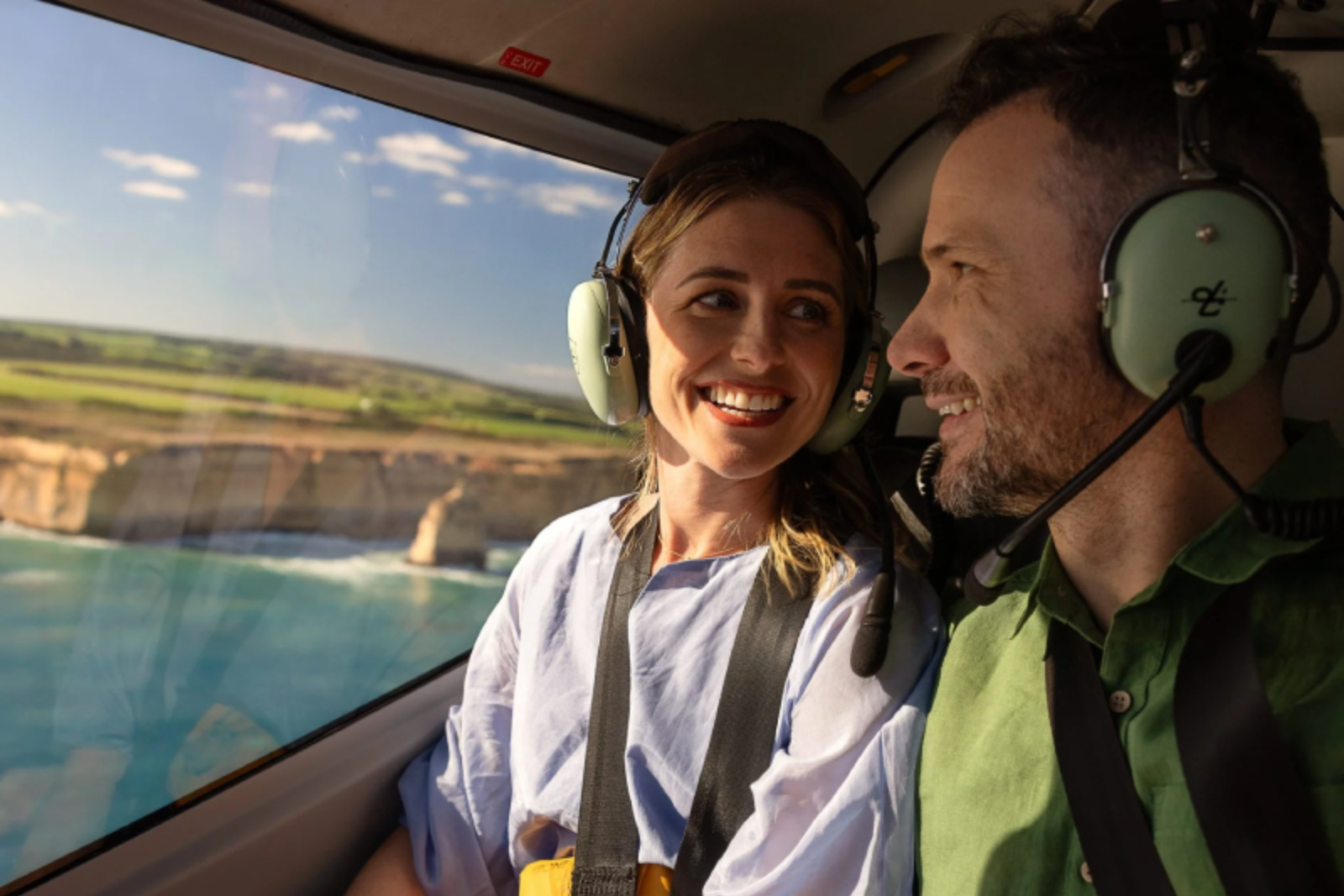 A couple in headsets smiles from inside a helicopter cockpit overlooking a coastal cliff landscape.
