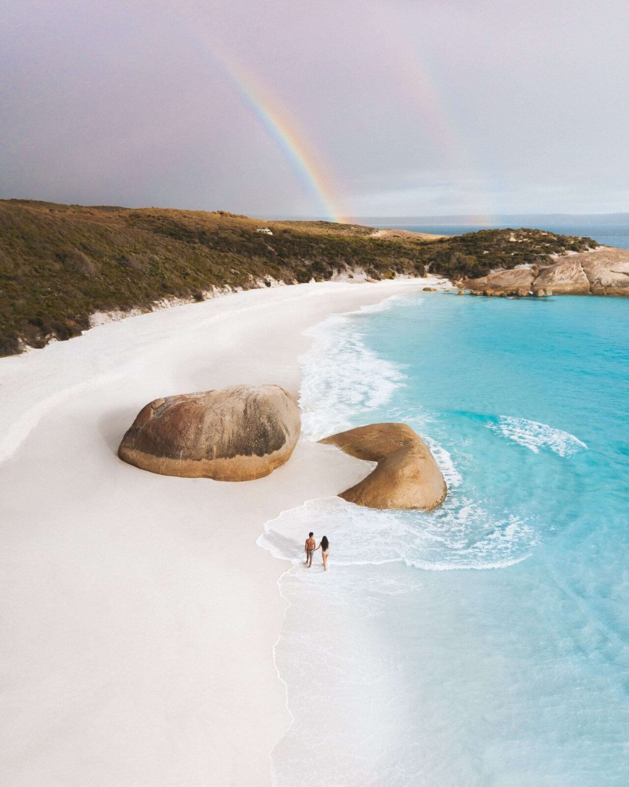 A rainbow arches over a secluded white-sand beach with turquoise water and pink granite boulders, with two people walking along the shoreline.