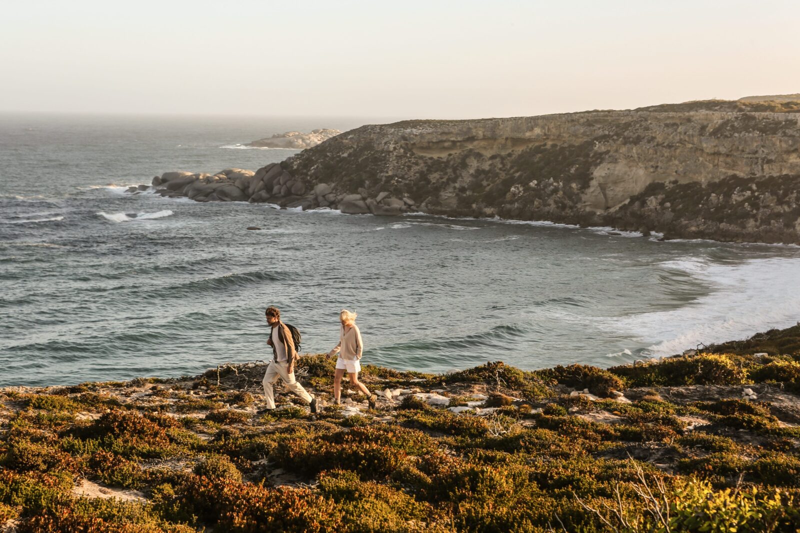 Two people walk along a rugged coastal cliff path above the ocean, surrounded by low green scrub.