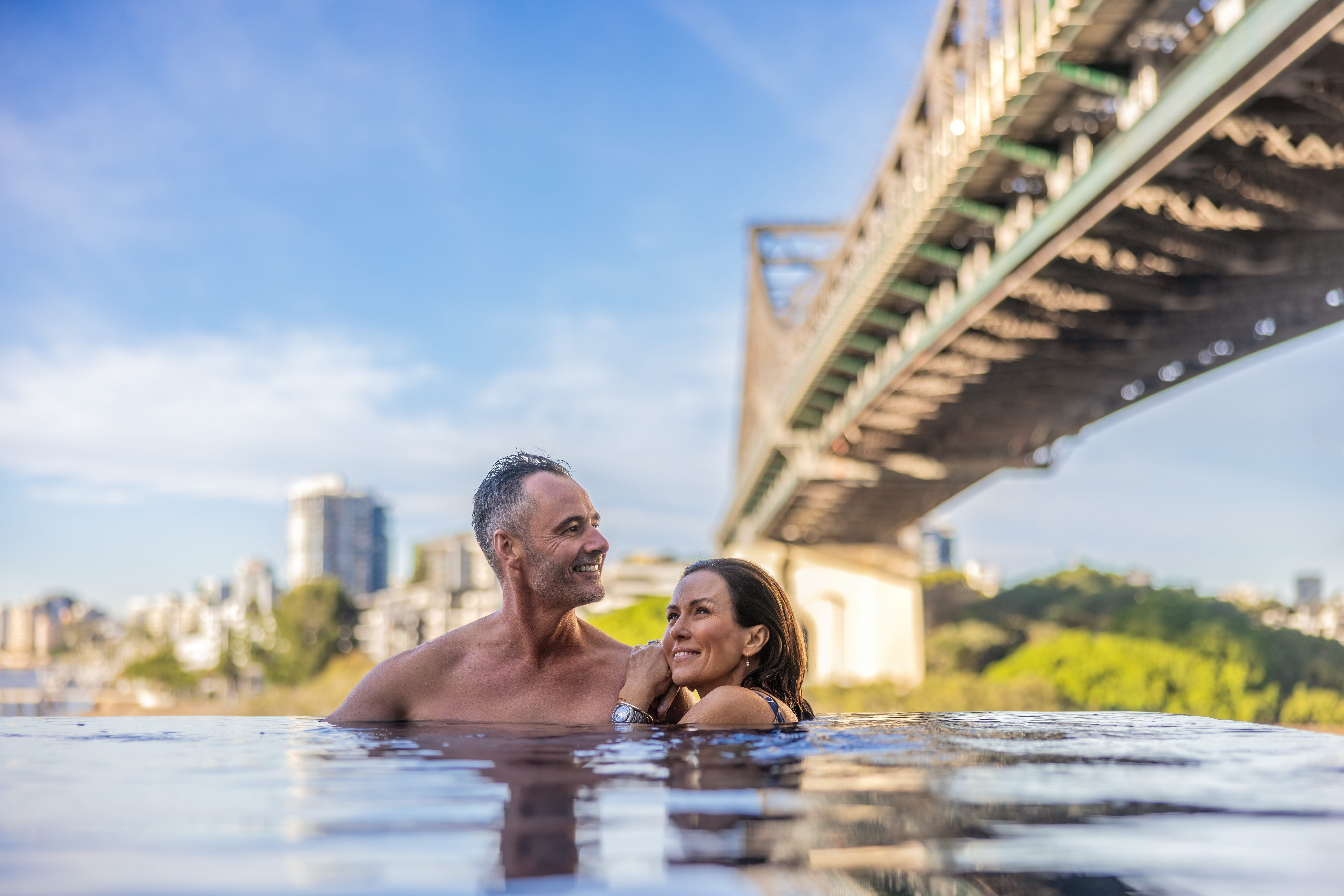 An older couple relax together in an infinity pool with a view of Sydney Harbour Bridge and the city skyline.