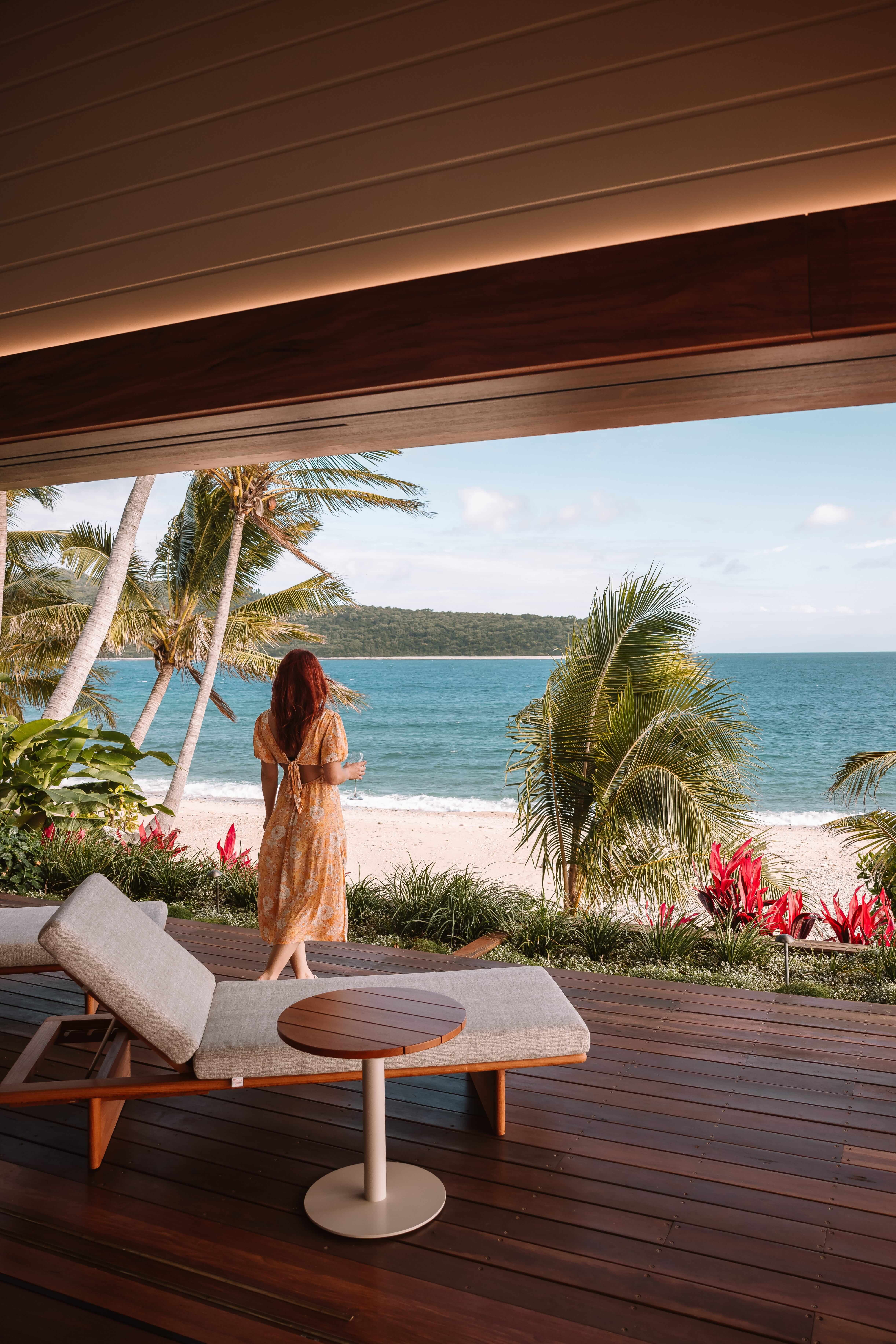 A woman in a flowing dress standing on a wooden deck, looking out at a tropical beach with palm trees and turquoise ocean.