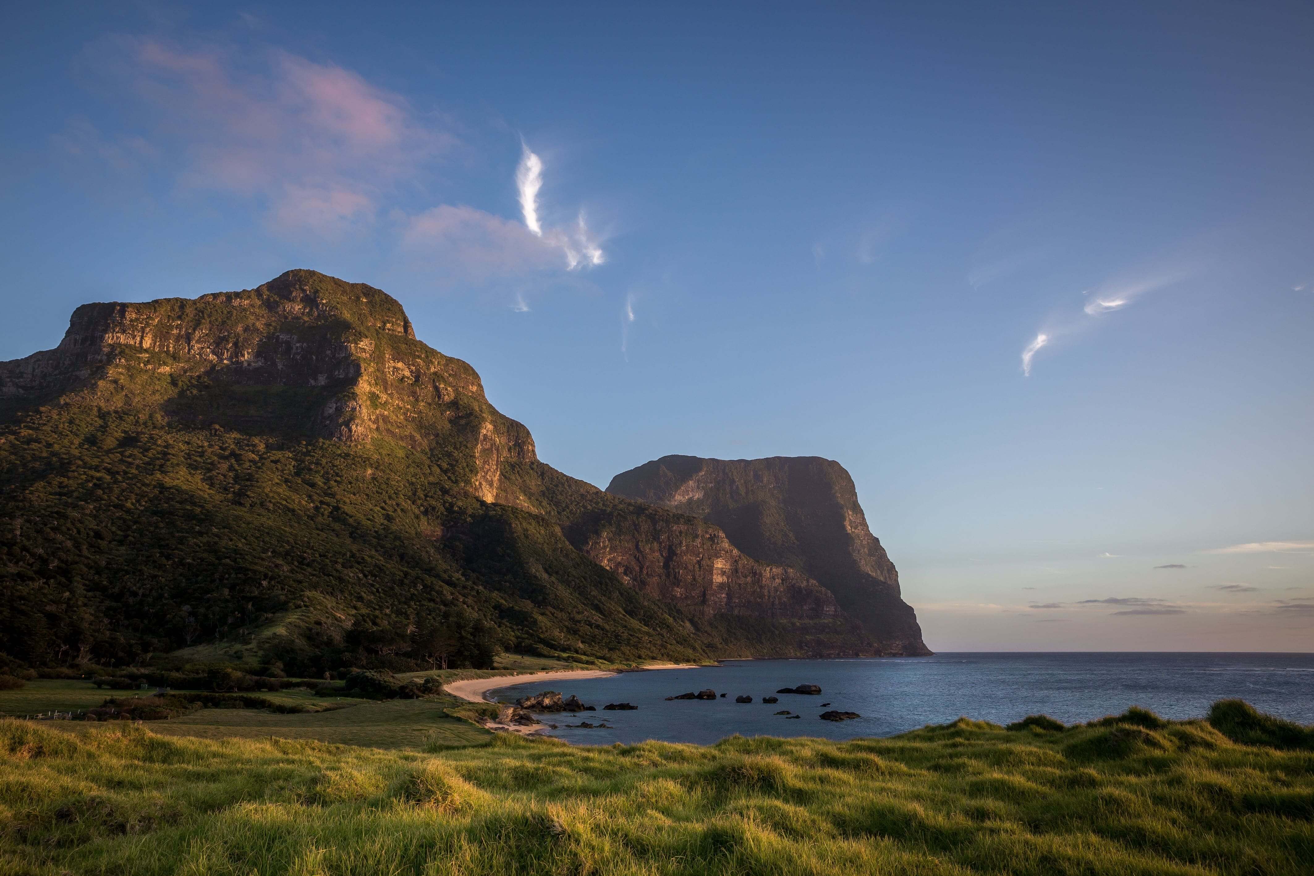 The twin peaks of Mount Gower and Mount Lidgbird rise above a calm bay and grassy headland on Lord Howe Island at dusk.