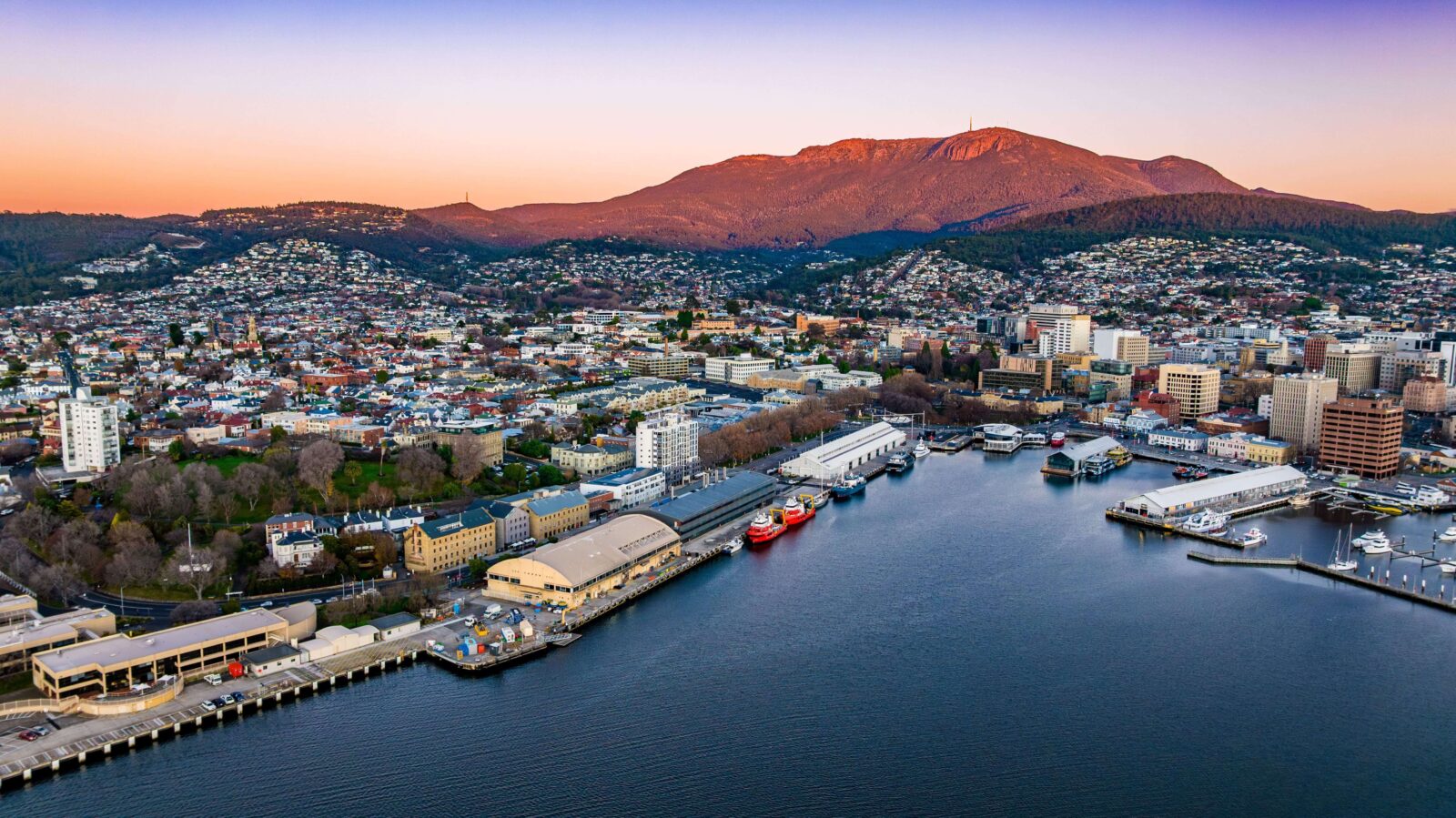 Hobart’s waterfront at dusk, with the harbour, historic wharf buildings, and kunanyi/Mount Wellington rising in the background.