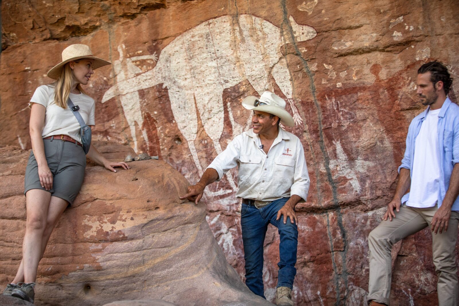 A guide points out ochre Aboriginal rock art on a red sandstone wall to visitors during an outback cultural tour. 