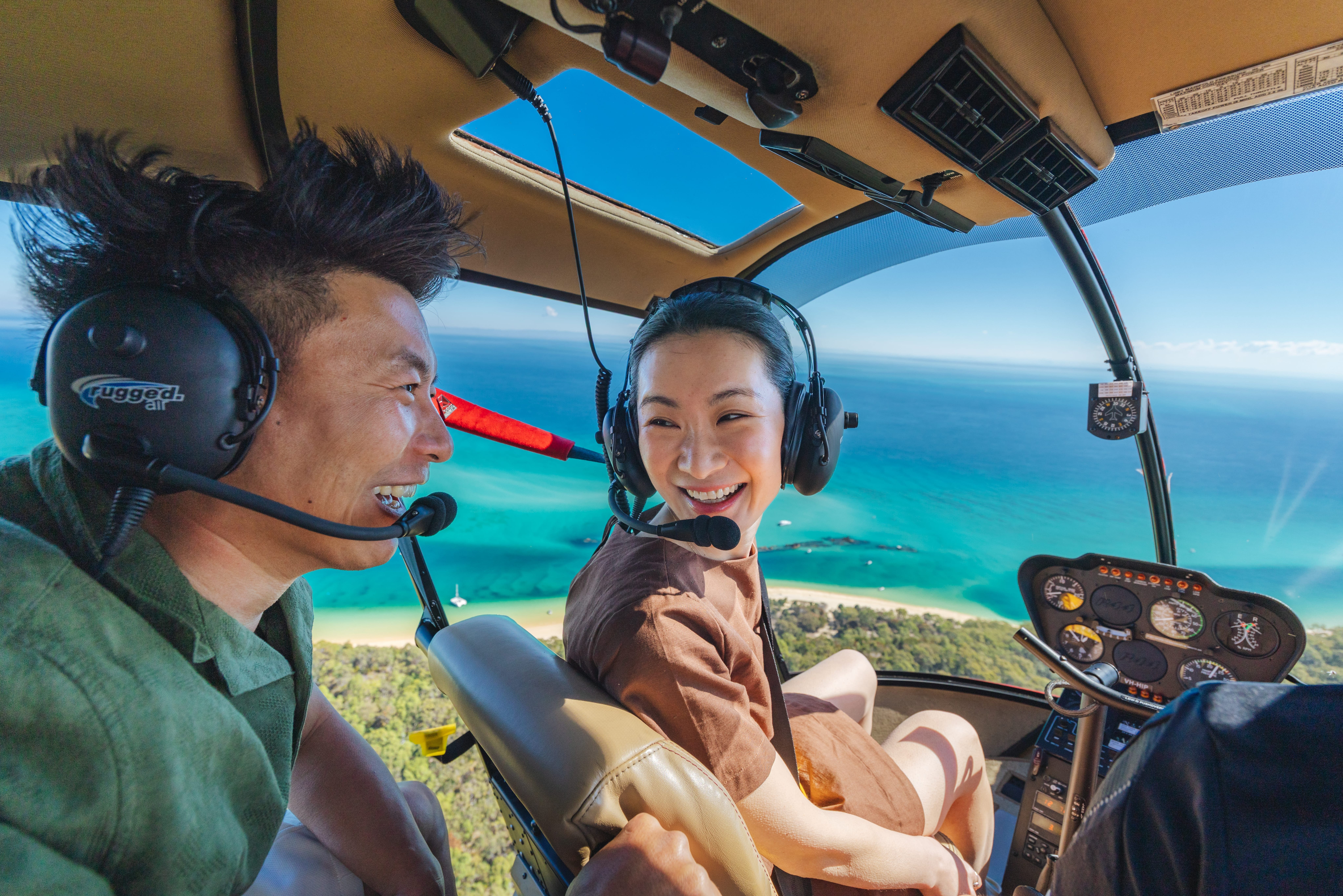 A couple wearing headsets smiles while riding in a helicopter over a turquoise coastline.