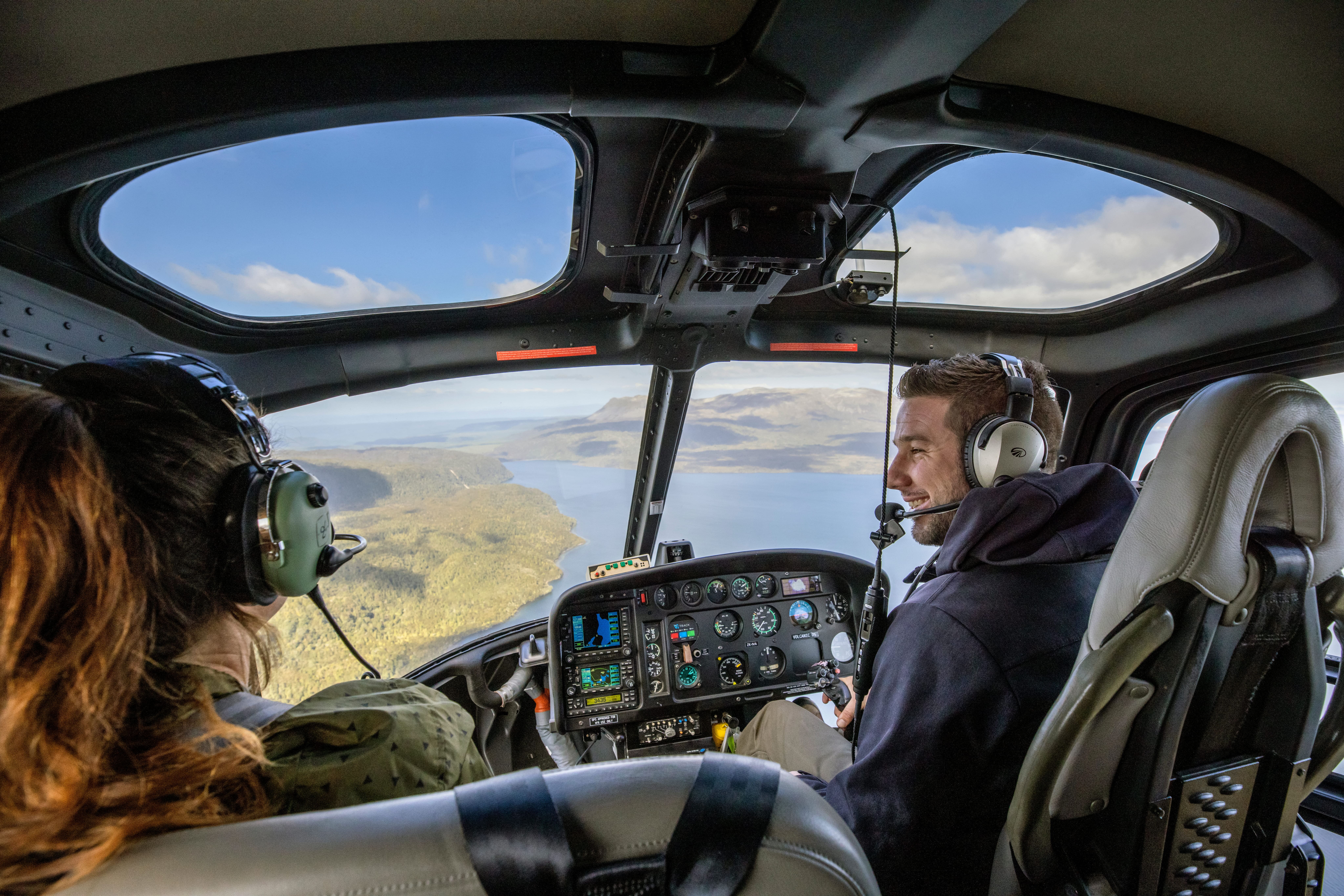 Two people wearing headsets inside a helicopter cockpit, flying over a coastal landscape  